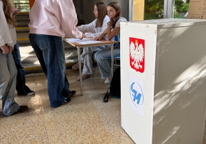 Uczennica podpisująca listę wyborczą na tle urny wyborczej.