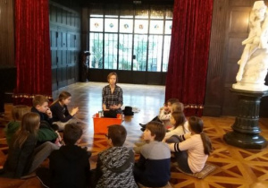 Uczniowie siedzą w kręgu i słuchają opowieści. Po prawej stronie znajduje się duża, biała rzeźba na czarnym postumencie, w oddali czerwone kotary i duże okno.