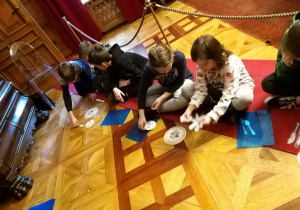 Uczniowie siedzą na czerwonym dywanie i uczą się prawidłowego nakrywania do stołu.