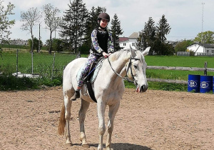 Dziewczynka ubrana w kask, kamizelkę ochronną i sztyblety siedzi na białym koniu.