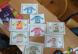 Dziewięć kart pracy ułożonych na ławce szkolnej z projektami swetrów świątecznych.