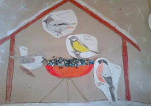 Praca plastyczna: karmnik dla ptaków. W karmniku wklejone zostały obazki: sikorki, wróbla, jemiołuszki i gila oraz ziarna stanowiace ptasi pokarm.