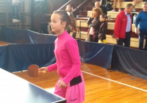 Reprezentantka szkoły w różowym dresie z rakietką w prawej ręce przy stole pingpongowym.