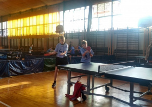 Dwóch zawodników w niebieskich koszulakach z logo szkoły w czasie meczu tenisa stołowego.