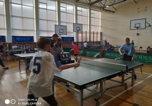 Dwóch zawodników w białej i niebiesce koszulce przy stole do tenisa stołowego rozgrywających mecz.