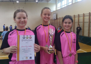 Szczęśliwe trzy uczennice w różowych koszulkach wraz ze zdobytymi medalami, dyplomem i pucharem.