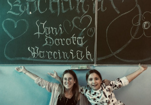 Dwie uśmiechnięte uczennice prezentujące napis wykonany na tablicy będący dedykacją dla nauczyciela plastyki.
