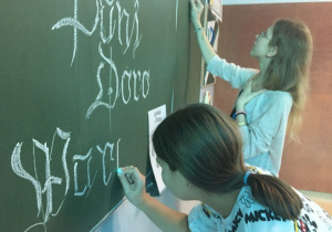 Dwie uczennice wykonujące napis białą kredą na zielonej tablicy.
