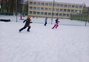 Troje uczniów na pierwszym planie biega po śniegu, kilku uczniow w oddali rzuca się śnieżkami.