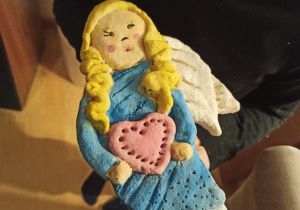 Aniołek ulepiony z masy solnej ubrany w niebieską sukienkę, W dłoniach trzyma duże, czerwone serce.