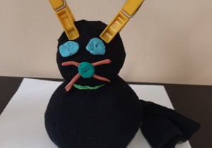 Maskotka przedstawiająca czarnego siedzącego kota. Kot ma oczy, nos i wąsy wkonane z plasteliny, a uszy ze spinaczy do bielizny.