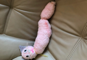 Maskotka przedstawiająca różowego leżącego kotka na kanapie. Kot ma szare uszka i żółtą wstążeczkę na szyi