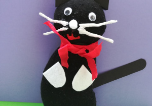 Maskotka przedstawiająca czarnego siedzącego kota. Kotek ma białe wąsy, czerwoną wstążeczkę zawieszoną na szyi.