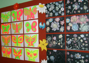 Wystawa prac uczniowskich: po lewej stronie znajduje się dwanaście prac, na których znajdują się na białym tle kolorowe motyle, a po prawej osiem prac ukazujących kwiaty namalowane kolorową kredą na ciemnym tle.