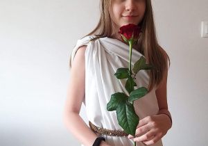 Dziewczynka ubrana w białą tunikę, w rękach trzyma różę, uczennica przebrała się za Afrodytę - boginię miłości, piękna i kwiatów.