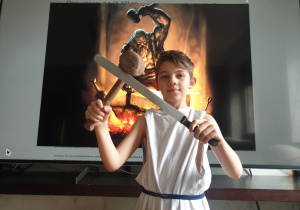 Chłopiec ubrany w białą tunikę, w prawej ręce trzyma młot, a w lewej nóż, miecz, w tle widać pracującego Hefajstosa - boga ognia i kowali.