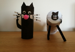 2.Dwa koty wykonane z rolek papieru: czarny kot, który stoi pionowo ma długie, białe w czarne paseczki wąsy i różowy nosek drugi biały ma czarną główkę i siedzi podparty na dwóch czarnych łapkach.