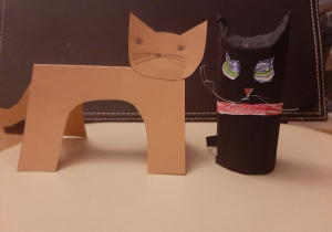 3.Dwa koty: czarny wykonany z rolki papieru i brązowy - wycięty z brystolu, stoi na czterech łapach. Czarny kot ma zielone oczy i czerwoną obróżkę.