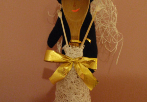 Kukiełka przypominająca elegancką kobietę, wykonana z drewnianej łyżki. Ubrana jest w czarną marynarkę i beżową koronkową spódnicę ozdobioną złotą kokardą. Na głowie ma czarny toczek z beżowym trenem.