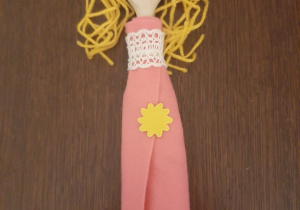 Kukiełka z drewnianej łyżki przedstawiająca młodą, złotowłosą dziewczynę. Ubrana jest w różową długą sukienkę, ozdobioną w górnej części białą koronką i żółtym kwiatkiem.