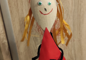 Kukiełka dziewczynki, wykonana z drewnianej łyżki. Ma złote włosy z błyszczących wstążeczek, narysowane uśmiechnięte usta i przyklejone oczy. Ubrana jest w czerwoną sukienkę uformowaną z papieru.