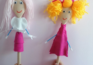 Kukiełki przedstawiające dwie dziewczynki, wykonane z drewnianych łyżek. Obydwie mają włosy z włóczki, uśmiechnięte buzie i przyklejone oczy. Jedna ubrana jest w różową sukienkę, a druga ma na sobie białą bluzkę z wacika i różową spódniczkę. Obydwie mają rączki z patyczków higienicznych.