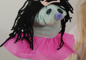 Pacynka dziewczynki w różowej spódniczce wykonana z niebieskiej skarpety. Na główce ma czarne oczy, niebieski nosek i czarne włosy z włóczki przypięte spinką.