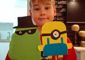 Chłopiec trzyma dwie kukiełki Minionków, wykonane z papieru i przymocowane do patyczków. Jedna postać jest dwuoka, zielona w czarnym kapeluszu i w czarnych okularach na oczach. Druga natomiast jednooka, żółta w niebieskich ogrodniczkach i w goglach na oku.