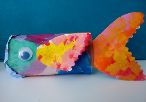 1.Kolorowa rybka na niebieskim tle wykonana z rolki papieru. Praca pomalowana jest farbami. Rybka ma doklejone niebieskie oczko oraz żółto-różowo-pomarańczowe płetwy i ogon.