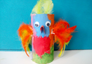 Kolorowa papuga wykonana z papierowej rolki, pomalowana wielobarwnie farbami. Z boku ma doklejone skrzydła z czerwonych i żółtych piór oraz żółty pióropusz na głowie.