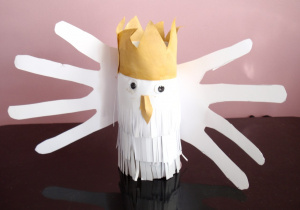 Biały ptak wykonany z papierowej rolki ze skrzydłami przypominającymi czteropalczaste dłonie. Tułów pokrywają paski naciętego papieru imitujące pióra. Na głowie ma założoną beżową koronę.