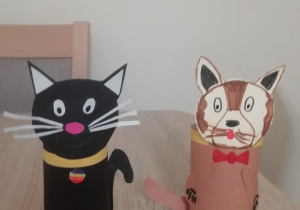 Dwa koty wykonane z rolek papieru w kolorach czarnym i brązowym. Oba maja doklejone głowy, łapki i ogonki. Czarny kot ma obróżkę z medalikiem, a brązowy z kokardą.