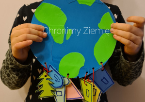 Dziecko trzyma w dłoniach wyciętą z papieru kulę ziemską. Do kuli przyczepione są na sznureczkach papierowe przywieszki: niebieska kropla, zielone drzewko oraz niebieskie, żółte, różowe i zielone trapezy symbolizujące kontenery na śmieci. Na kuli ziemskiej zanajduje się napis "Chronimy przyrodę".