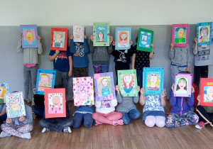 Zdjęcie uczniów klasy 2b trzymających prace plastyczne przedstawiające narysowany kredkami portret mamy.