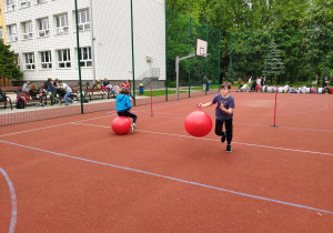 Zawodnicy podczas wyścigu rzędów: jedna uczennica skacze na piłce uszatce, a drugi uczeń wraca biegiem do zespołu trzymając piłkę w ręku.