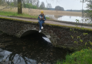 Uczennica czyta książkę siedząc nad wodą na kamiennym mostku.