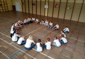 Duża grupa uczniów siedzi w kole wraz z nauczycielem w sali gimanstycznej. Wszyscy są ubrani w białe t-shirty i dżinsowe spodnie.
