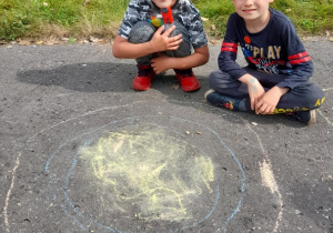 Dwóch uśmiechniętych chłopców z klasy 2b. Jeden z chłopców kuca, a drugi siedzi w siadzie skrzyżnym na asfaltowym boisku przy wykonanym przez siebie rysunku wielkiej kropki.