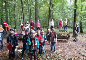 Zdjęcie przedstawia uczniów klasy 1a podczas zajęć na terenie Lasu Łagiewnickiego. Częśc uczniów pozuje do zdjęcia stojąc na ziemi, kilkoro dzieci stoi na pniu powalonego drzewa.