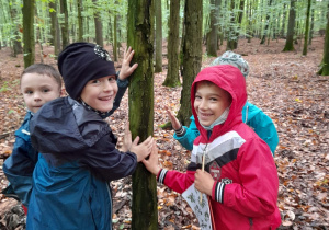 Zdjęcie przedstawia uczniów klasy 1a podczas zajęć na terenie Lasu Łagiewnickiego. Czterech uczniów dotyka ręką pnia drzewa.