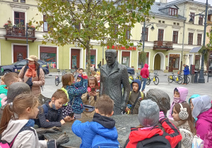 Uczniowie oglądają Pomnik Trzech Fabrykantów: I. Poznańskiego, K. Scheiblera i H. Grohmana . Siadają na wolnych miejscach przy stole . W tle widać jedną z kamienic przy ul. Piotrkowskiej.