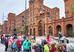 Uczestnicy wycieczki stoją przed dawną bramą fabryki Izraela Poznańskiego. Słuchają Pani Przewodnik.