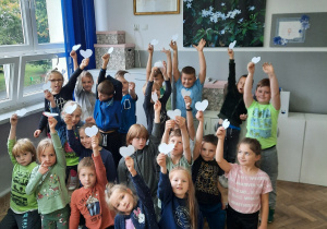 Uczniowie klasy 1a z białymi serduszkami w dłoniach stoją przed obrazem Świętego Jana Pawła II.