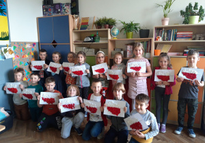 Zdjęcie przedstawia uczniów klasy 1a w pracowni lekcyjnej, którzy prezentują swoje prace wykonane z okazji Święta Niepodległości - kontur Polski wypełniony barwami narodowymi, z wykorzystaniem białej i czerwonej bibuły karbowanej formowanej w kształt kuleczek.