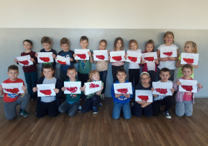 Zdjęcie przedstawia uczniów klasy 1a na korytarzu szkolnym, którzy prezentują swoje prace wykonane z okazji Święta Niepodległości - kontur Polski wypełniony barwami narodowymi, z wykorzystaniem białej i czerwonej bibuły karbowanej formowanej w kształt kuleczek.