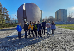Grupa uczniów stoi przed pomnikiem Pękniętego Serca, w oddali widać betonowy parkan z nazwami miejscowości, w których istniały inne obozy zagłady.