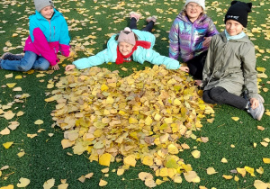 Cztery uśmiechnięte dziewczynki w kolorowych jesiennych kurtkach i czapkach. Trzy z nich siedzą na zielonej części boiska szkolnego, jedna leży. Przed dziewczynkami znajduje się ułożone z liści duże, żółte serce.