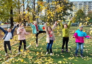 Sześć uśmiechniętych dziewczynek podrzuca do góry żółte liście. Większość z nich ma uniesione do góry ręce, jedna podnosi lewą nogę w bok. W tle widać zieloną część boiska szkolnego i jesienne drzewa.