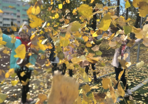 Na pierwszym planie zdjęcia widzimy wiele żółtych jesiennych liści podrzuconych przez uśmiechnięte dziewczynki, które delikatnie można dojrzeć na drugim planie.