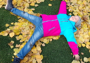 Dziewczynka w dżinsowych spodniach, różowo- błękitnej kurtce i szarej czapce leży na zielonym boisku szkolnym i wykonuje „Aniołka” wśród żółtych, jesiennych liści.